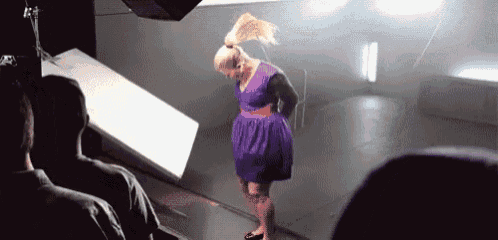 a woman in a purple dress is standing in front of a group of people in a dark room .