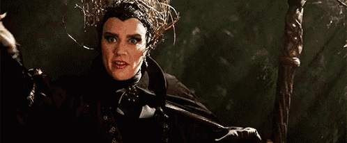 a woman in a black costume with a crown on her head is standing in the woods holding a sword .