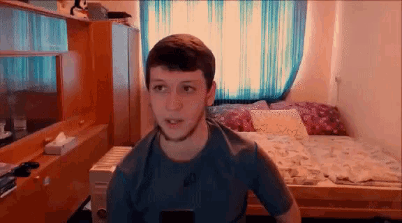 a man in a blue shirt is sitting in front of a bed and looking at the camera .