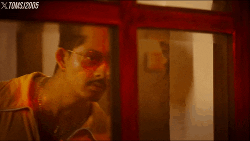 a man wearing glasses and a yellow jacket is looking through a red door with xtoms / 2005 in the corner