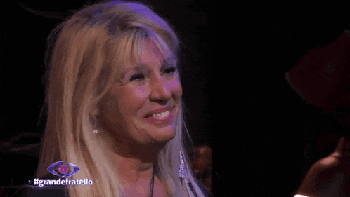 a woman with blonde hair is smiling in a dark room with #grandefratello written on the bottom