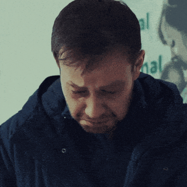 a man in a blue jacket looks down with a sign in the background that says hal
