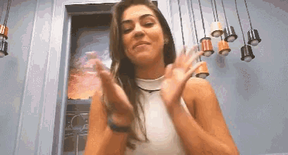 a woman in a white tank top is clapping her hands in front of a wall with hanging lights .