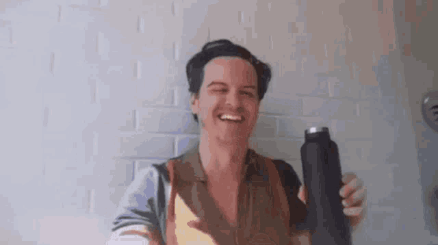 a man is smiling while holding a black bottle in front of a brick wall .