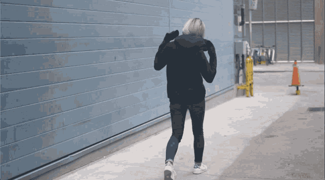 a woman in a black jacket is walking down a sidewalk in front of a blue wall