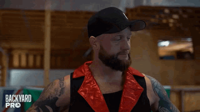 a man with a beard and tattoos is wearing a black hat and a red vest that says backyard pro