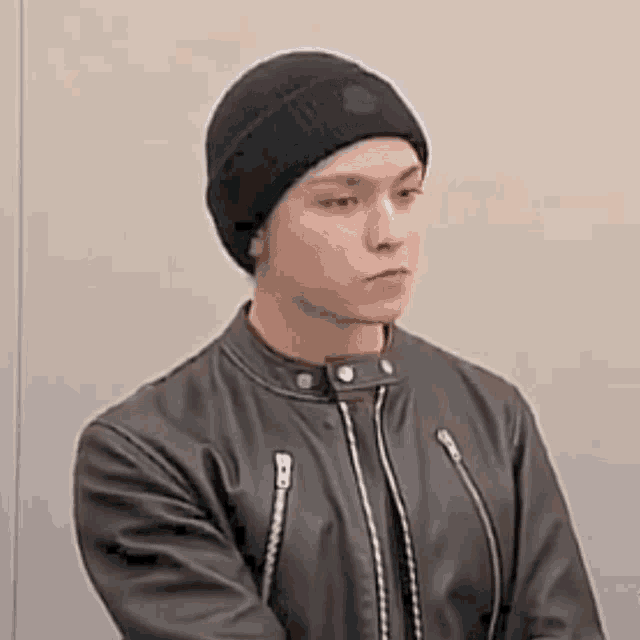 a young man wearing a black beanie and a leather jacket is standing in front of a wall .
