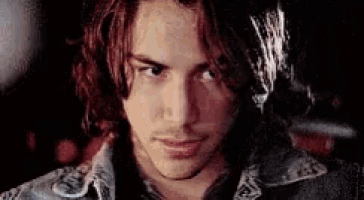 a close up of a young man with long hair wearing a denim jacket and looking at the camera .