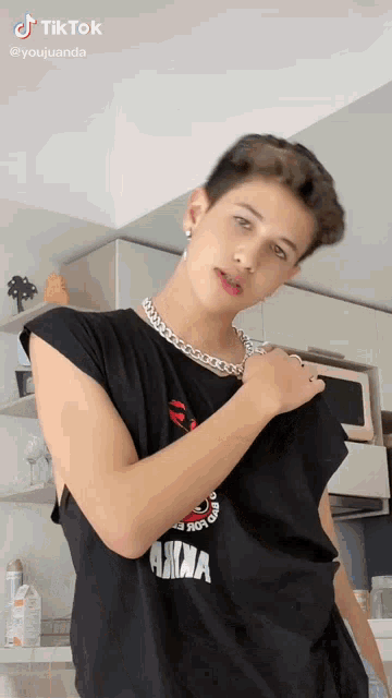 a young man wearing a black t-shirt and a chain around his neck is standing in a kitchen .