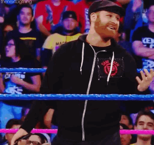 a man in a black hoodie is standing in a wrestling ring talking to the crowd .