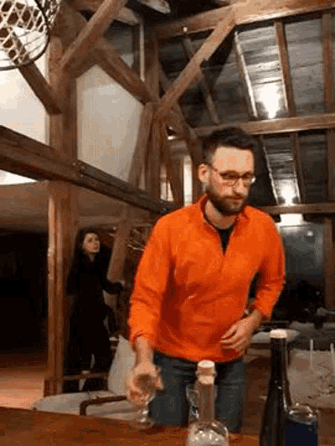 a man in an orange shirt is pouring a glass of wine from a bottle on a table .