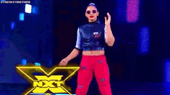 a woman in a crop top and red pants is standing on a stage in front of a sign that says nxt .