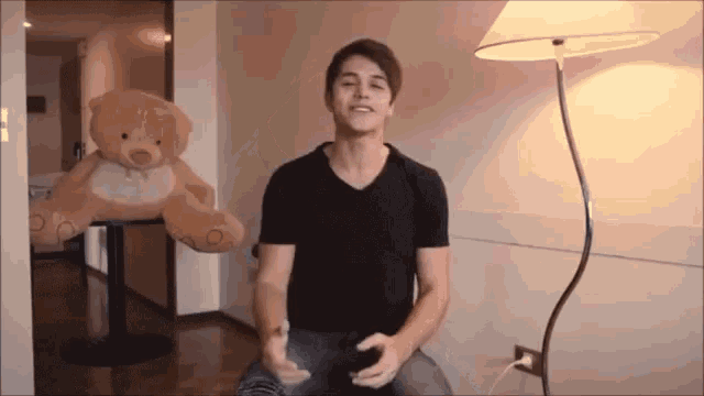 a man in a black shirt is sitting in front of a teddy bear and a lamp
