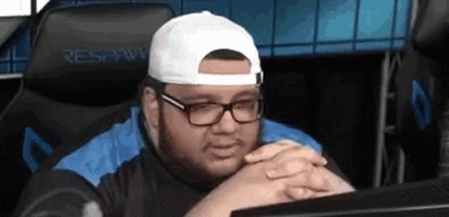 a man wearing glasses and a white hat is sitting in a chair in front of a computer screen .