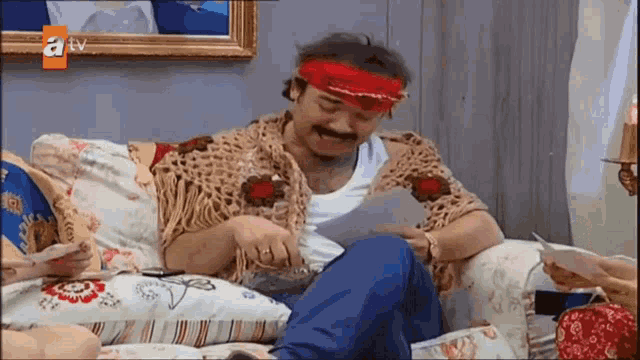 a man is sitting on a couch reading a piece of paper with a tv logo in the corner