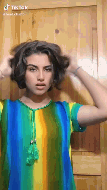 a woman in a rainbow colored shirt is holding her hair in front of a wooden wall with tiktok written on the bottom right