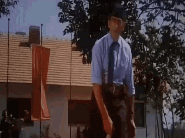 a man in a blue shirt and tie is standing in front of a house with flags hanging from the roof .