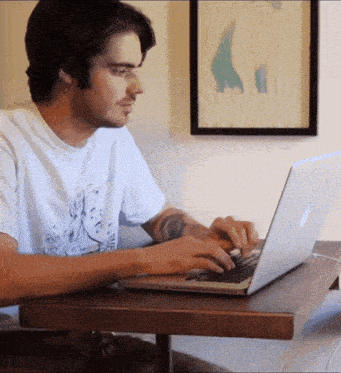 a man is typing on an apple laptop while wearing a white shirt that says the devil