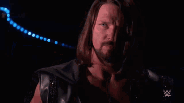 a man with long hair and a beard is standing in a dark room with a wrestling logo in the corner