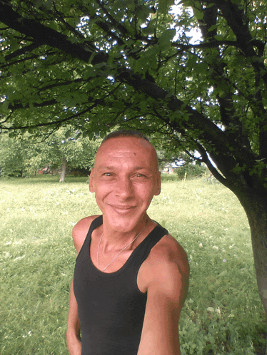 a man in a black tank top stands under a tree in a field