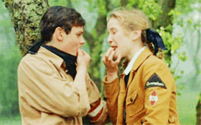 a boy and a girl are standing next to each other and the girl is wearing a jacket with a red cross on it