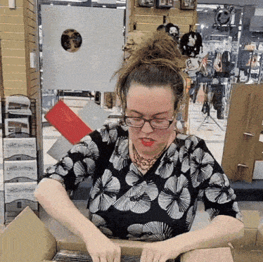 a woman wearing glasses and a black and white floral shirt is opening a box