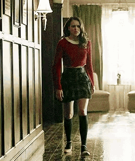 a woman is walking down a hallway wearing a red sweater and a plaid skirt