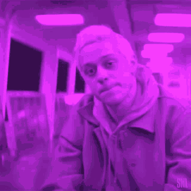 a man in a purple jacket is sitting in a dark room with a snl logo in the corner .