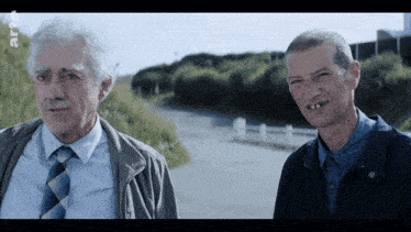 two men are standing next to each other on a road and one of them has a missing tooth