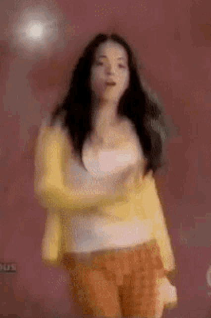 a woman in a yellow jacket and orange shorts is dancing in front of a pink wall
