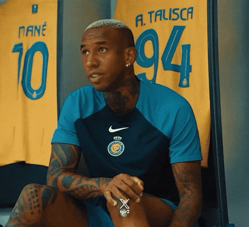 a man in a blue nike shirt sits in front of a yellow a.talisca jersey