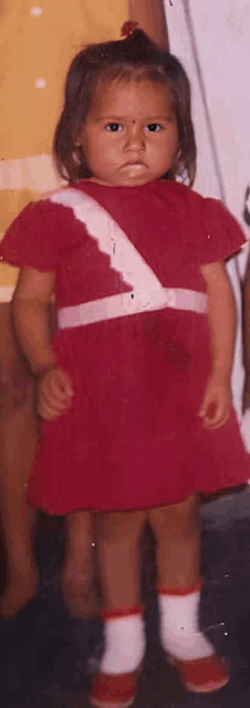 a little girl in a red dress and white socks stands in front of a yellow shirt