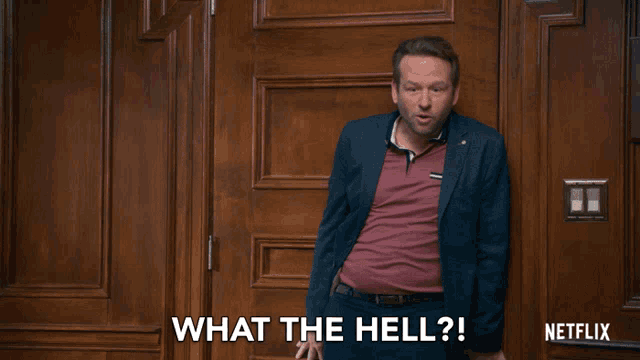 a man in a suit is standing in front of a wooden door and says what the hell