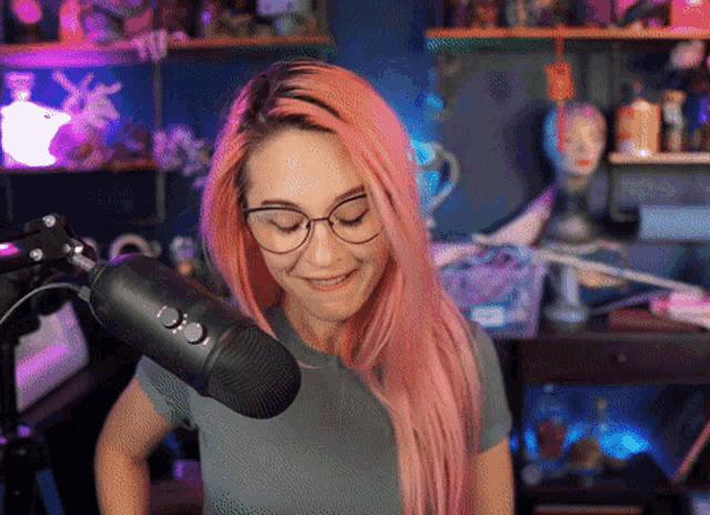 a woman with pink hair and glasses stands in front of a black microphone