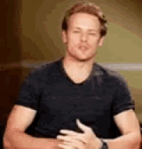 a man in a black shirt is sitting with his hands folded in front of him .