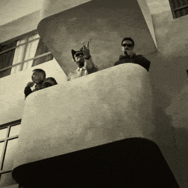 three men are standing on a balcony and one of them is wearing sunglasses
