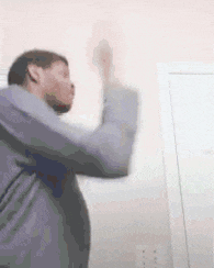 a man in a gray shirt is standing in front of a door and holding his hands up in the air .