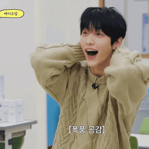 a young man wearing a tan sweater with korean writing on the bottom right corner