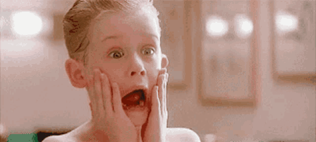 a young boy with a surprised look on his face is holding his hands to his face .