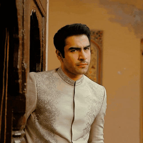 a man in a white shirt with silver embroidery on the collar stands in front of a door