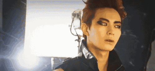 a young man with red hair and black eye makeup is wearing a black vest and looking at the camera .