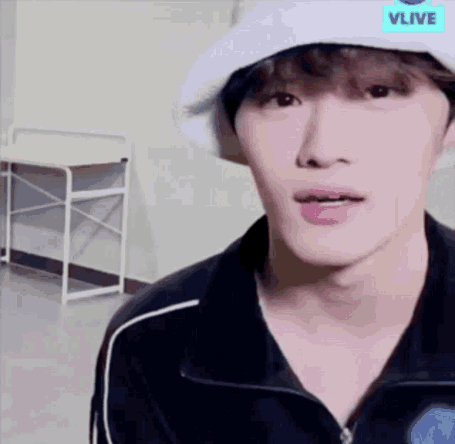 a young man wearing a white hat and a black jacket is standing in front of a desk .