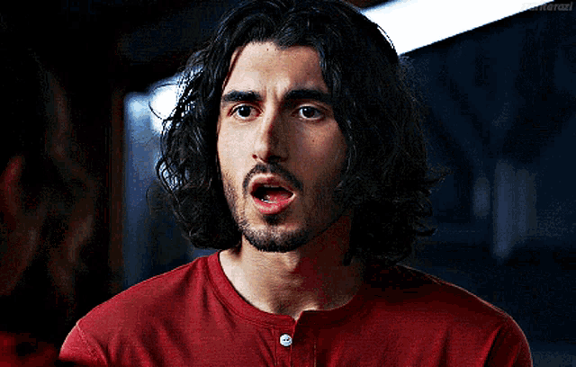 a man with long curly hair and a beard is wearing a red shirt .