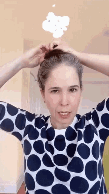 a woman wearing a blue and white polka dot shirt is holding her hair in a bun
