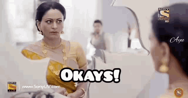a woman in a yellow saree is looking at herself in a mirror and says " okays "