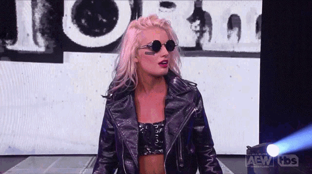 a woman wearing sunglasses and a leather jacket is standing in front of a screen that says aew tbs .