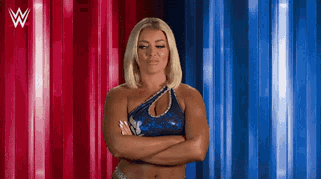 a female wrestler is standing with her arms crossed in front of a red and blue curtain .