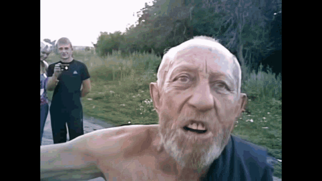 a shirtless man with a beard looks at the camera while another man takes a picture of him