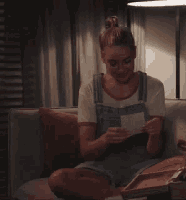 a woman in overalls is sitting on a couch reading a letter