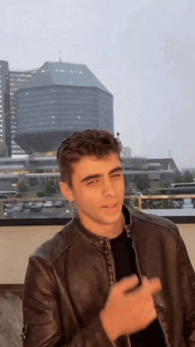 a young man wearing a leather jacket is standing on a rooftop in front of a city skyline .